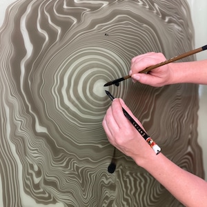 May include: An abstract artwork featuring a concentric, tree-ring-like design in shades of brown and white. Two hands hold calligraphy brushes, applying ink to the surface, creating a unique artistic composition.