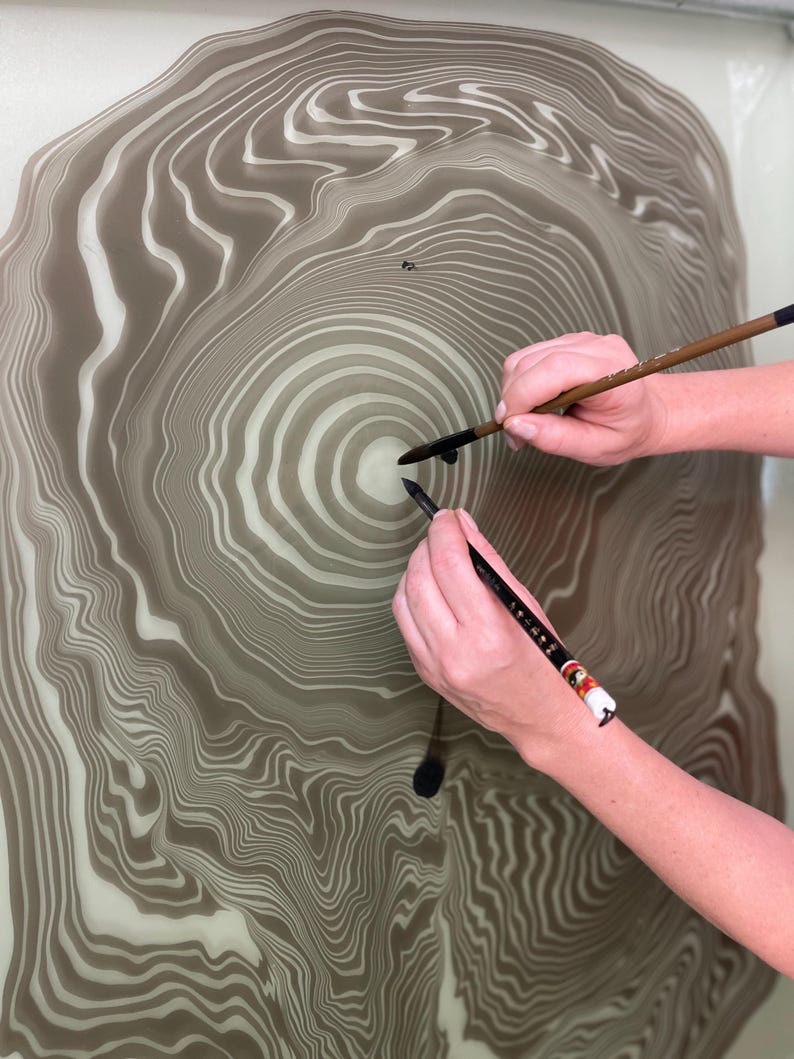 May include: An abstract artwork featuring a concentric, tree-ring-like design in shades of brown and white. Two hands hold calligraphy brushes, applying ink to the surface, creating a unique artistic composition.