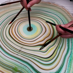 May include: A close-up of a colorful abstract art piece with concentric circles in shades of green, brown, yellow, and white. Several paintbrushes are positioned over the artwork, suggesting the process of creation. The artwork has a marbled effect.