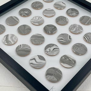 May include: A framed artwork featuring numerous circular, gray and white marbled designs. The circles are arranged on a white background within a black frame. The marbled patterns vary, creating a unique visual texture.