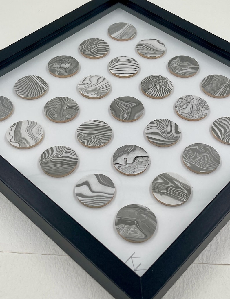 May include: A framed artwork featuring numerous circular, gray and white marbled designs. The circles are arranged on a white background within a black frame. The marbled patterns vary, creating a unique visual texture.