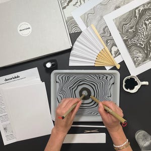 Puede incluir: Un kit de arte de marmoleado con tinta flotante. Incluye una bandeja con un diseño en blanco y negro, tinta, pinceles, papel, un abanico y ejemplos impresos. El kit es de decoriolab.