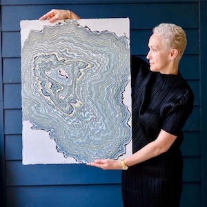 May include: A rectangular piece of textured paper featuring an intricate, swirling marbled pattern in shades of light blue, sage green, cream, and dark charcoal. The design resembles topographical lines or geological strata on a white background.