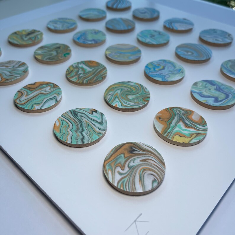 May include: A collection of round, wooden coasters with a swirled, marbled design. The coasters feature shades of teal, orange, yellow, and white. They are arranged on a white surface, creating a visually appealing pattern. The coasters are ideal for home decor.