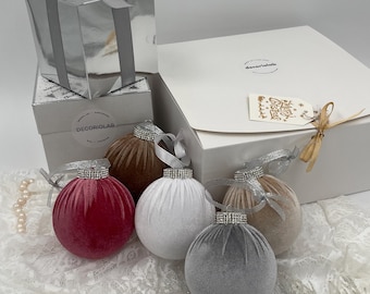 Puede incluir: Una colección de adornos navideños de terciopelo en rojo, blanco, marrón, gris y beige, cada uno con una tapa con incrustaciones de diamantes de imitación y una cinta plateada. Los adornos se exhiben con cajas de regalo y una etiqueta que dice "Felices Fiestas".