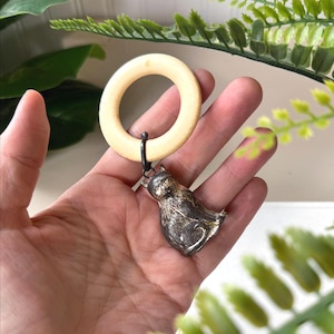 May include: A cream-colored wooden ring with a small, detailed silver cat charm attached. The cat charm is in a sitting position. The ring is connected to the charm by a small black metal loop.