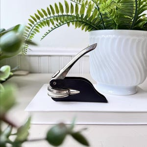 May include: A vintage black and silver seal press, likely for embossing. The press features a long, curved silver handle and a black base. The seal is resting on a stack of white books, with a white ceramic planter and greenery in the background.