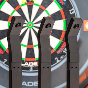 Peut inclure: Trois supports de caméra de jeu de fléchettes noirs avec câbles attachés sont placés devant une cible de fléchettes. La cible a un cadre noir et un motif cible coloré. Les supports sont sur une surface en bois.