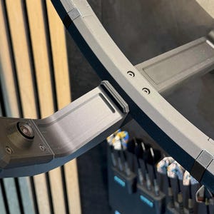 May include: A close-up of a black camera mounted on a curved, gray metal frame. The frame is attached to a larger, curved structure with visible screws. The background features vertical wooden slats and a glimpse of golf clubs.