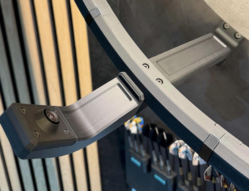 May include: A close-up of a black camera mounted on a curved, gray metal frame. The frame is attached to a larger, curved structure with visible screws. The background features vertical wooden slats and a glimpse of golf clubs.