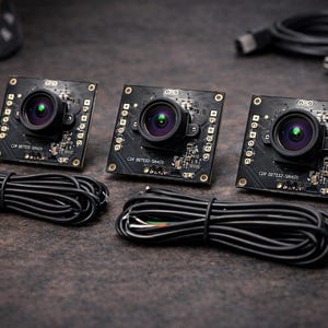 May include: Three black square circuit board cameras with lenses are arranged on a dark surface. Each camera has a lens with a green reflection. Black cables and a USB cable are also present. A dartboard is visible in the background.
