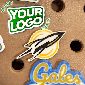 Pode incluir: Uma coleção de amuletos de sapatos coloridos. Os amuletos incluem um design "YOUR LOGO" verde e branco, um foguete e um design "Gales" azul e amarelo. Os amuletos estão em uma superfície marrom.