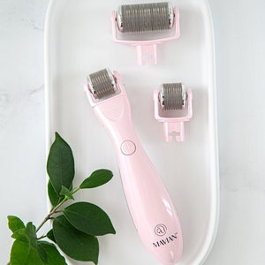 May include: Three pink and silver facial rollers on a white oval plate. The largest roller has a pink handle with the word "MAVIAN" printed on it. A small green plant is in the background.