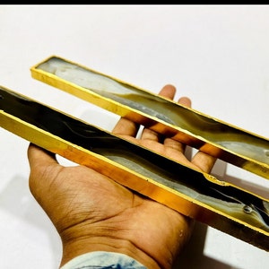 May include: Two rectangular decorative pieces with gold-coloured metal frames. The interior features a swirling pattern of grey, black, and white. The pieces are held in a person's hand against a white background.