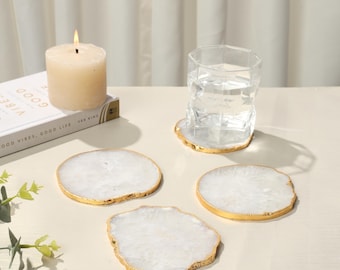 May include: Four white agate coasters with gold trim. The coasters are round and have an irregular shape. They are on a white surface.