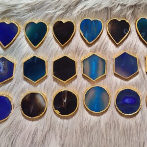 May include: A collection of decorative coasters in various shapes: hearts, hexagons, and circles. Each coaster features a polished agate stone in shades of blue, teal, and black, with a gold-colored metallic trim. The coasters are arranged on a fluffy, light-colored surface.