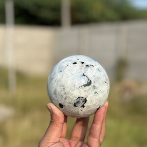May include: A white and black moonstone sphere with iridescent rainbow flashes. The sphere is being held in a hand.