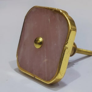 May include: A square pink and gold cabinet knob with a gold screw. The knob is made of a natural stone with a gold metal frame.
