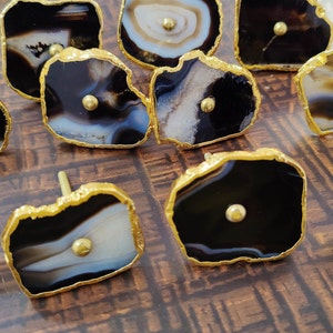 May include: A set of 11 agate drawer pulls with gold trim. The agate is black and white with a natural pattern. The pulls are all different shapes and sizes.
