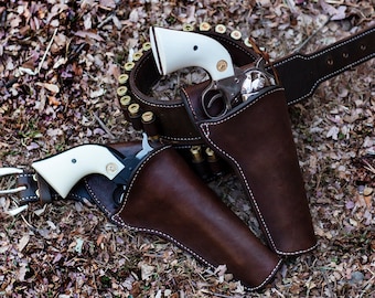 May include: Two brown leather holsters with white-handled revolvers, attached to a brown leather belt with a row of brass bullets.