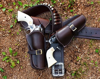 May include: Two brown leather holsters with silver buckles and ivory grips attached to a brown leather belt with silver buckle and bullet loops.