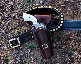 May include: A brown leather gun holster with a silver revolver and a brown leather belt with a silver buckle and a row of bullets.