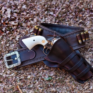 Può includere: Una cintura per pistola in pelle marrone con una fibbia argentata e una fondina. La cintura ha una fila di cartucce in ottone attaccate ad essa. La fondina è vuota.