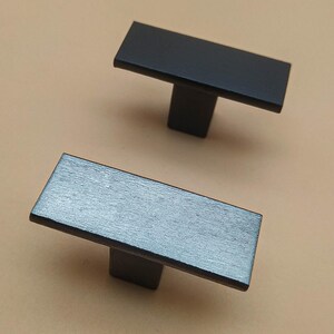 May include: Two rectangular cabinet pulls. One is black, the other is a gray-toned wood grain. Each pull has a rectangular handle attached to a square base. The pulls are set against a neutral background.