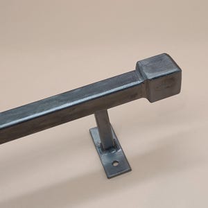 Könnte beinhalten: Eine dunkelgraue Metallstange mit quadratischem Ende und Halterung. Die Stange ist rechteckig und scheint aus Stahl zu sein. Die Halterung ist an der Stange befestigt und hat ein einzelnes Loch zur Montage. Der Hintergrund ist hellbeige.