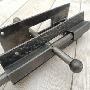 May include: A close-up of a dark grey, hammered metal door bolt. The bolt has a rectangular design with a sliding bar and a round handle. The metal has a textured surface and is mounted on a light wood surface.