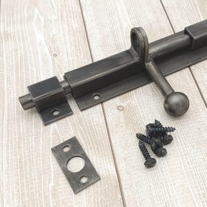 May include: A dark grey metal sliding bolt latch with a round handle and mounting hardware. The latch is shown with a strike plate and screws, suitable for doors or gates. The hardware is displayed on a light wood grain surface.
