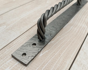 May include: A black metal door pull with a twisted rope design. The pull is mounted on a rectangular plate with holes for screws.