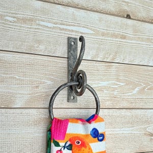 Può includere: Un anello portasciugamani e un gancio in metallo forgiato a mano montati su una parete di legno. Il gancio presenta un design a spirale decorativo. Un asciugamano colorato con un motivo a pappagallo è appeso all'anello.