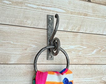 May include: A hand-forged metal towel ring and hook fixed to a wooden wall. The hook has a decorative swirl design. A colourful towel with a parrot design hangs from the ring.