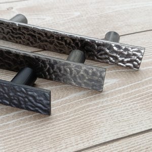 Könnte beinhalten: Nahaufnahme von zwei Metall-Schrankgriffen mit gehammerter Textur. Die Griffe sind rechteckig mit zylindrischem Griff. Einer ist dunkelgrau, der andere hellgrau. Der Hintergrund ist eine helle Holzmaserung.