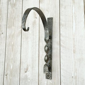 May include: A dark gray, hand-forged metal hanging basket bracket. The bracket features a curved hook and a decorative, twisted central support. The mounting plate has a textured surface and is designed for wall mounting. The bracket is made of iron.