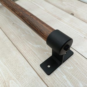 Puede incluir: Una manija de puerta de metal negro con una barra de madera. La manija está montada en una pared con un soporte de metal negro.