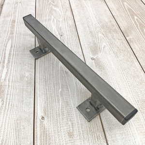 Peut inclure: Une rampe d'escalier rectangulaire en métal gris avec deux plaques de montage carrées. La rampe est constituée d'un tube carré épais et est conçue pour une prise en main robuste. Le métal a une finition industrielle brute.