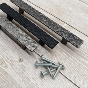 May include: Four metal cabinet pulls with a hammered texture in silver, black, and gray. The pulls are rectangular with rounded ends and are accompanied by a pile of silver screws. The hardware is displayed on a light wood surface.