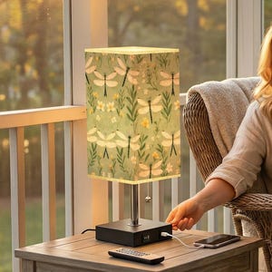 May include: A table lamp with a dragonfly pattern on the shade sits on a small wooden table. The lamp is next to a woman sitting in a wicker chair on a porch. The text "PORCH CALM meets MODERN CONVENIENCE" is at the bottom of the image.