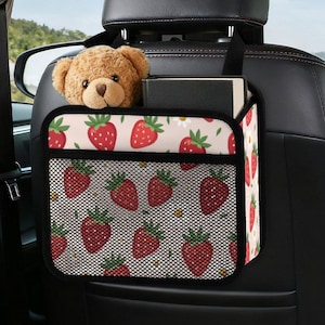 Puede incluir: Un organizador de asiento de coche con un estampado de fresas, colgado del reposacabezas. El organizador tiene un bolsillo de malla negro y está lleno de un oso de peluche y libros. El fondo muestra el interior de un coche.