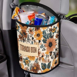 May include: A floral patterned trash bin with a black zippered top and the words "Trash Bin" printed on the front. The bin is hanging on the back of a car seat.
