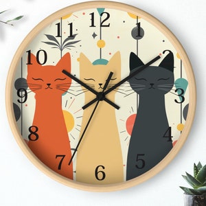 May include: A round wall clock with a white background and black numbers. The clock features three stylized cats with closed eyes, one orange, one yellow, and one black. The clock has a wooden frame.