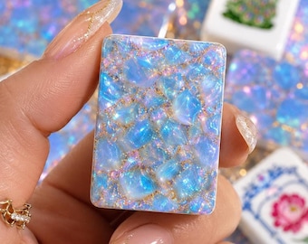 Könnte beinhalten: Ein rechteckiger Mahjong-Spielstein mit einer mosaikartigen Oberfläche aus schillernden blauen und hellvioletten steinartigen Fragmenten, die in eine goldene Glitzerharzbasis eingebettet sind und eine texturierte Optik erzeugen.