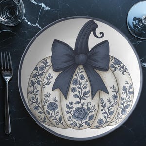 May include: A decorative plate featuring a pumpkin design with a large bow and floral accents. The pumpkin is cream-colored with a dark blue bow and stem. The plate is decorated with blue floral patterns and is perfect for fall decor.