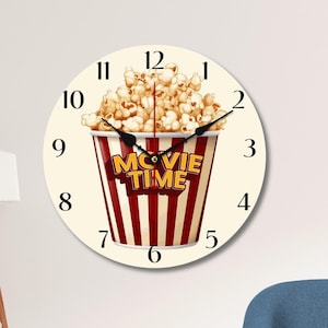 Peut inclure: Une horloge ronde blanche avec des chiffres et des aiguilles noirs. Le cadran de l'horloge présente un seau de popcorn rayé rouge et blanc avec les mots "Movie Time" en lettres jaunes.