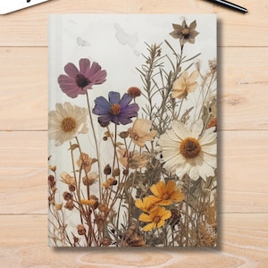May include: A white journal with a pressed flower design. The flowers are in shades of purple, yellow, and white. The journal is on a wooden surface.