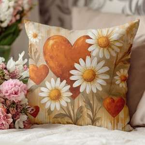 Puede incluir: Un cojín decorativo con un diseño de corazón y margaritas. El cojín tiene un fondo crema con corazones naranjas y margaritas blancas con centros amarillos. El cojín es de forma cuadrada y está colocado sobre una superficie blanca.