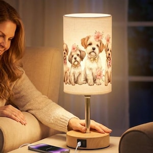 May include: A decorative table lamp with a wooden base and a cylindrical lampshade featuring a print of several dogs with pink flowers. The lamp has a gold-colored stem and a USB charging port. A woman is sitting next to the lamp.