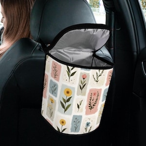 May include: A car trash bag with a floral pattern. The bag is white with rectangular blocks of pastel colors, each featuring a different flower illustration. The bag hangs from the headrest and has a black zipper and interior.